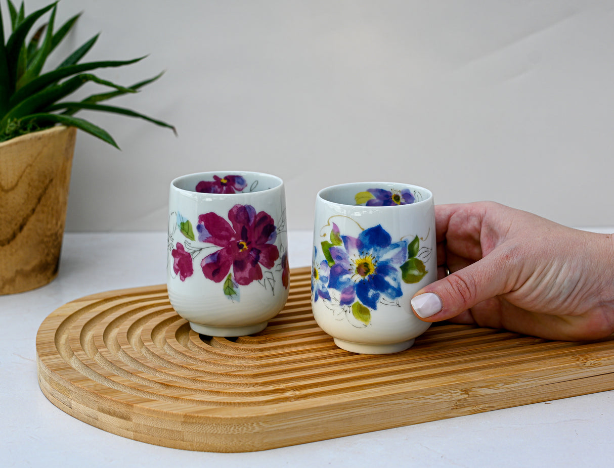 A hand cradles a Pink Flower Waisted Tea Cup, crafted with traditional Japanese ceramic techniques, alongside another exquisite piece on a wooden tray. A verdant plant graces the background, completing this delicate tableau.