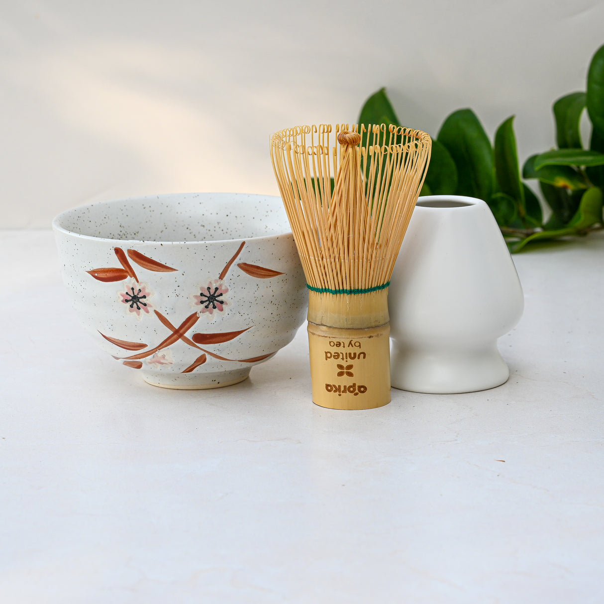 A Bamboo Matcha Bowl, Whisk, and Holder Set rests elegantly on a light surface with a white vase, while green leaves create a serene backdrop.
