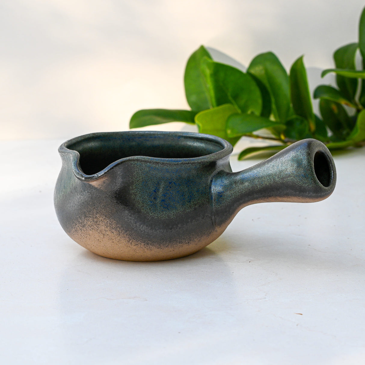 The Blue Spouted Ceramic Matcha Bowl, handcrafted in Japan, features a spout and handle. It sits on a white surface, framed by green leaves in the background.