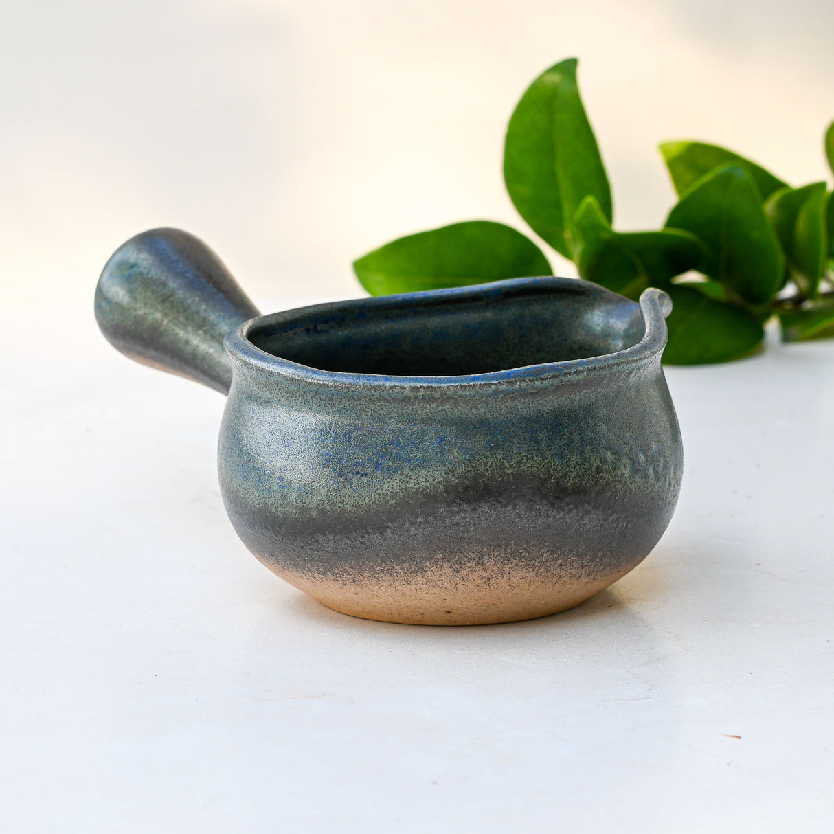 A Blue Matcha Bowl with Spout sits on a white surface, with green leaves in the background.