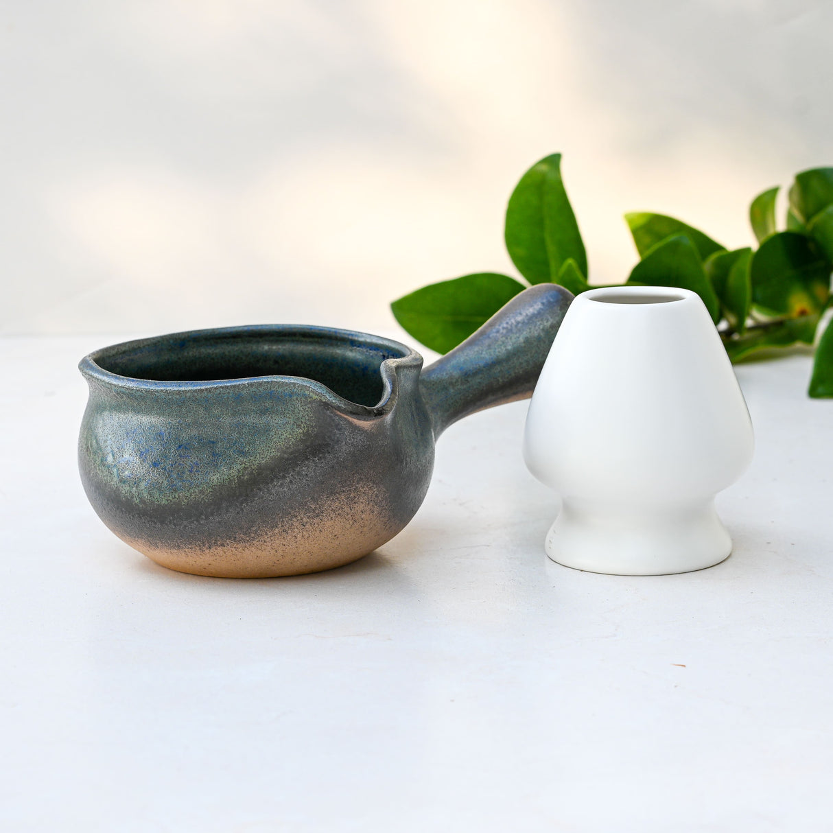 Blue Matcha Bowl with Spout next to a white cup; green leaves in the background on a light surface.