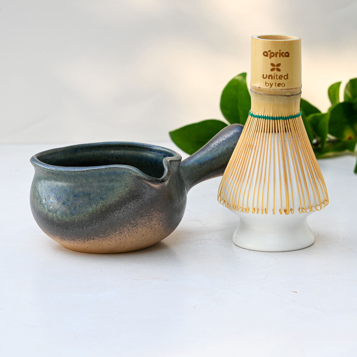 A Blue Matcha Bowl with Spout and a bamboo whisk rest on a white surface, green leaves in the background, highlighting timeless Minoyaki ceramic artistry.