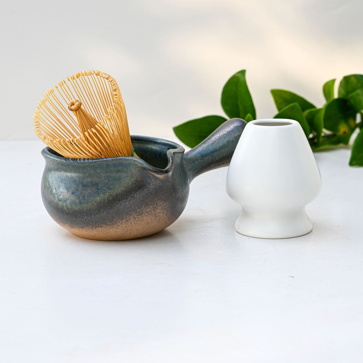 The Blue Matcha Bowl with Spout sits beside a bamboo whisk, white cup, and green leaves on a light surface.