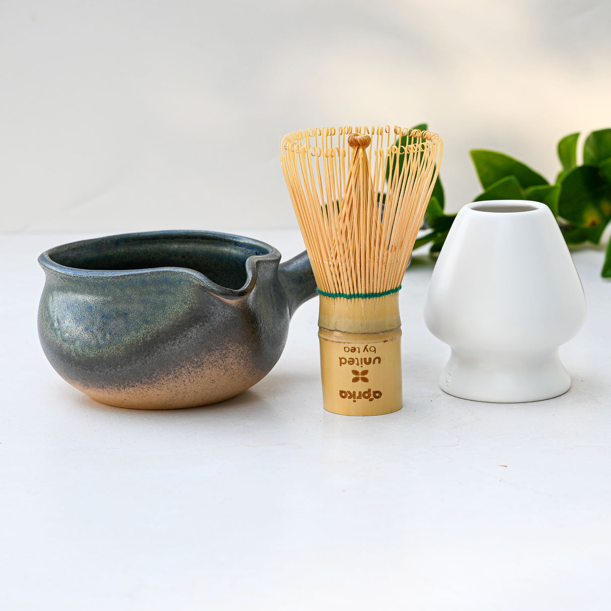 Blue Matcha Bowl with Spout, bamboo whisk, and white vase arranged on a light surface with green leaves in the background. This Japanese handcrafted bowl adds elegance and tradition to your tea ritual.