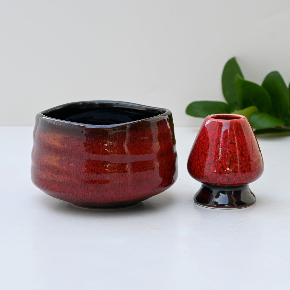 A Red Matcha Bowl with a glossy finish sits elegantly on a light surface, reminiscent of Minoyaki Pottery. Nearby, a green plant adds serenity, evoking the tranquility of traditional Matcha Tea Ceremonies.