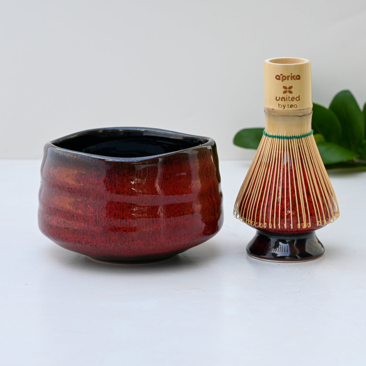 A Red Matcha Bowl and bamboo tea whisk sit on a light surface among green leaves, capturing the elegance of traditional matcha tea ceremonies.