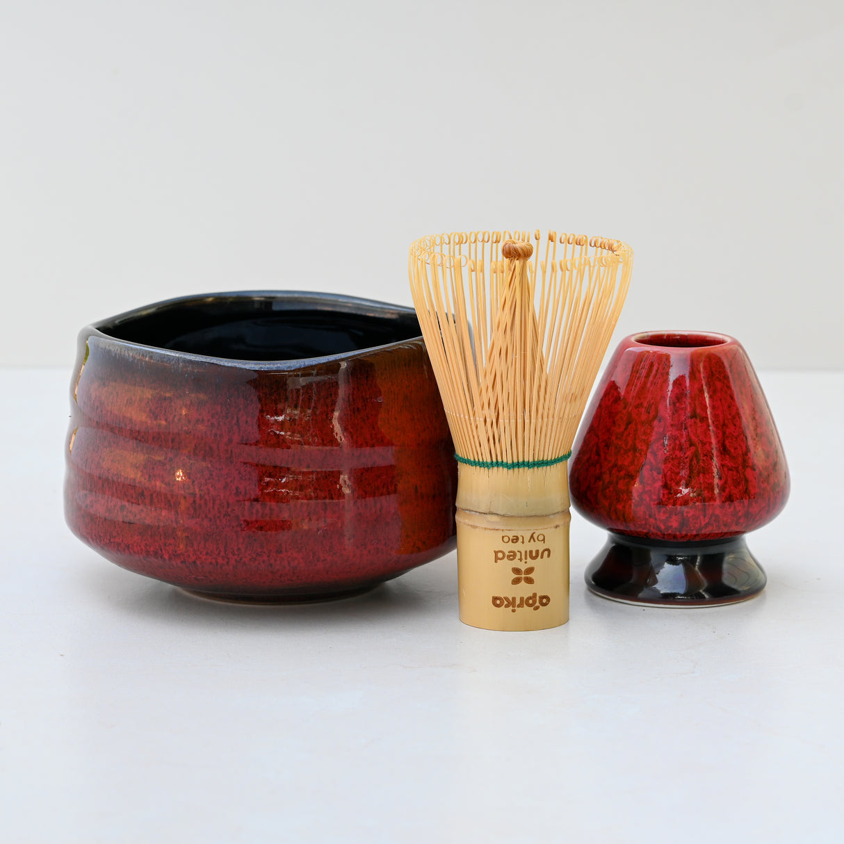 A Red Matcha Bowl, paired with a bamboo whisk and holder, along with a matching red stand, rest on a light surface, capturing the serene elegance of traditional Matcha tea ceremonies.