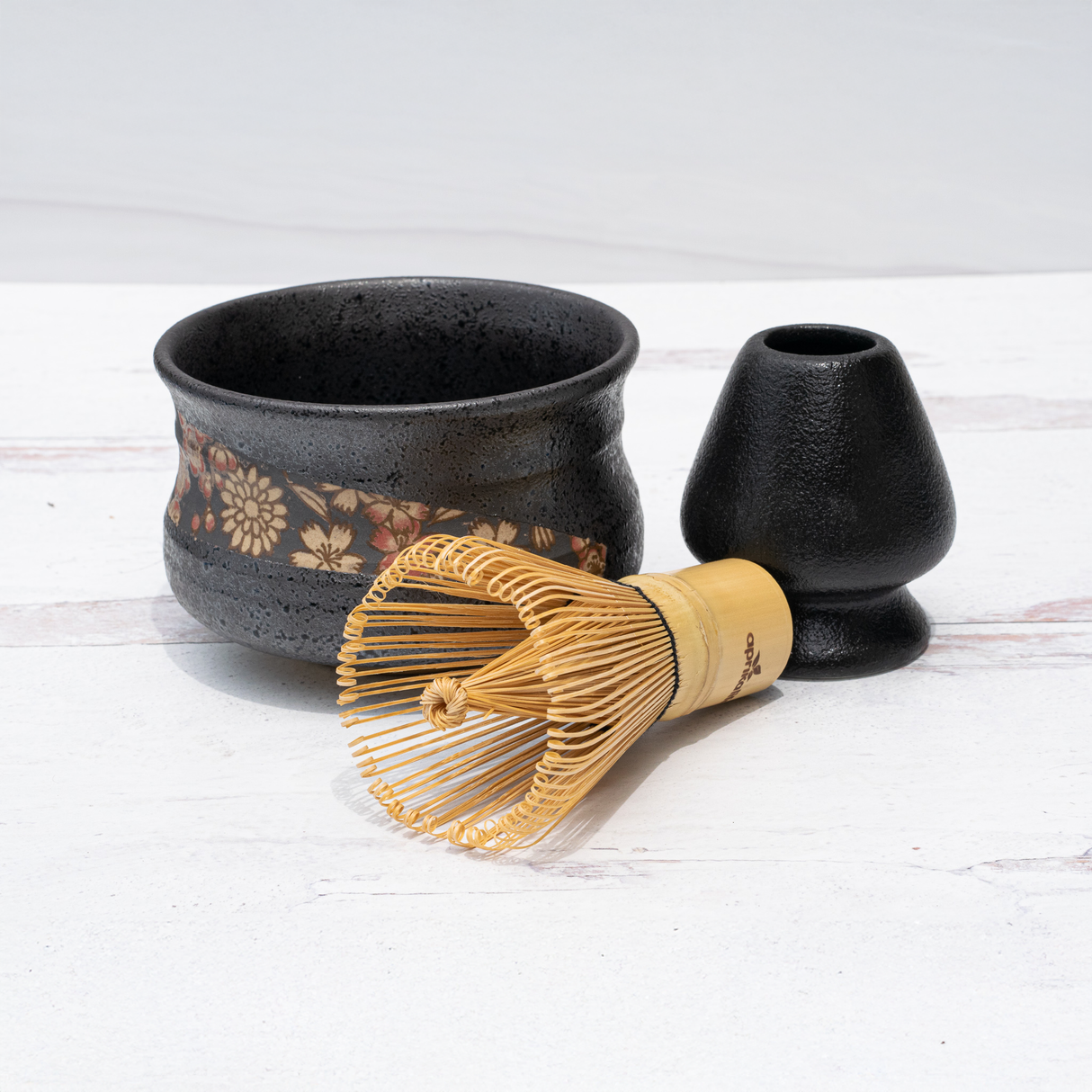 A Black with Flowers Matcha Bowl and whisk holder complement a bamboo matcha whisk on a white surface.