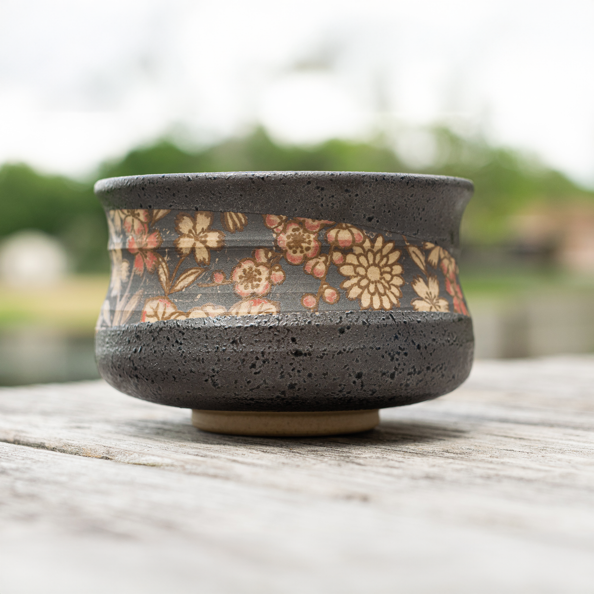 The Black with Flowers Set, a ceramic matcha bowl adorned with floral patterns and accompanied by a bamboo whisk, rests on a wooden table outdoors against a blurred natural backdrop.