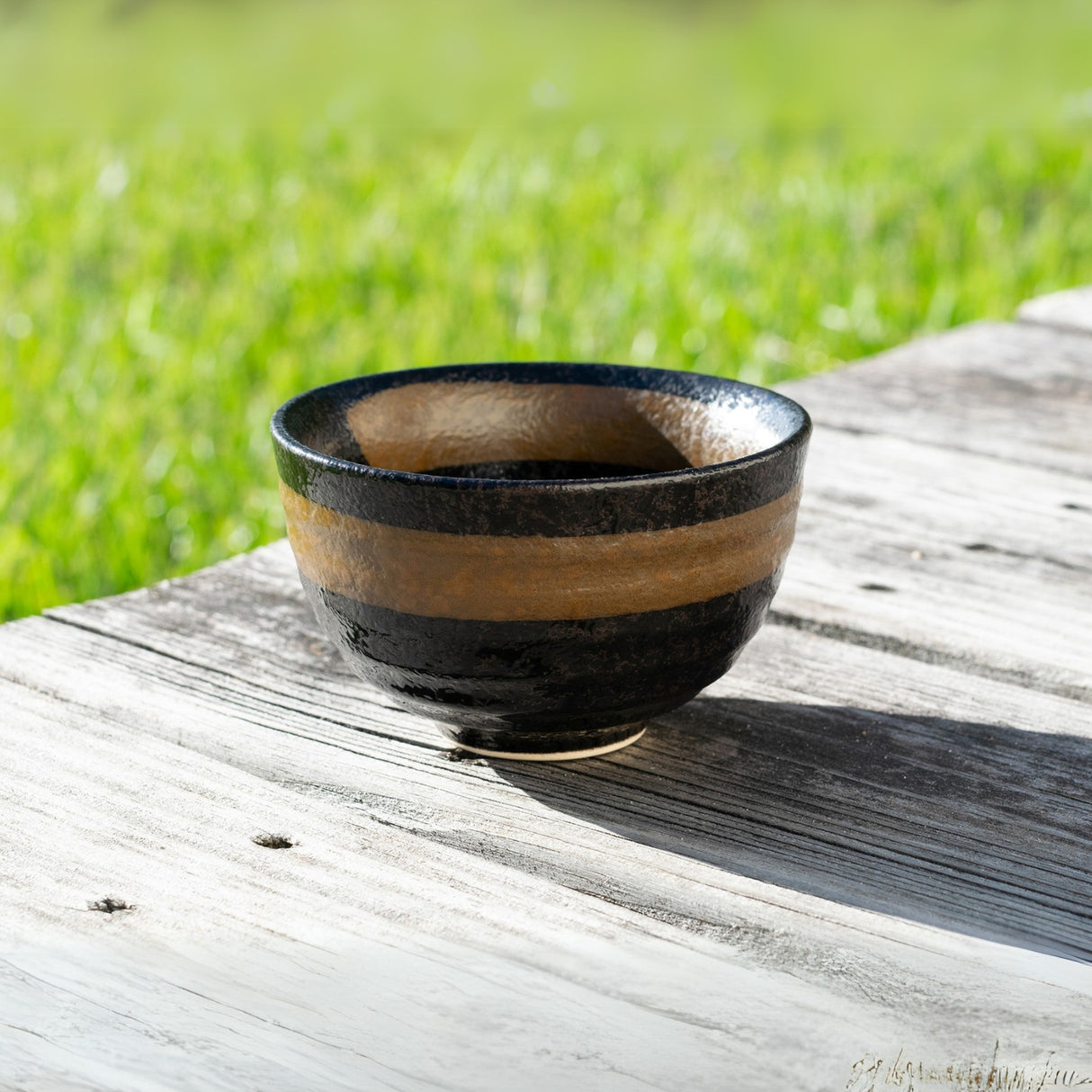 A Dark Brown Glaze Ceramic Matcha Bowl, part of the set with a bamboo whisk, whisk holder, and organic Japanese matcha, crafted using Minoyaki pottery techniques, rests on a weathered wooden table against a grassy background.