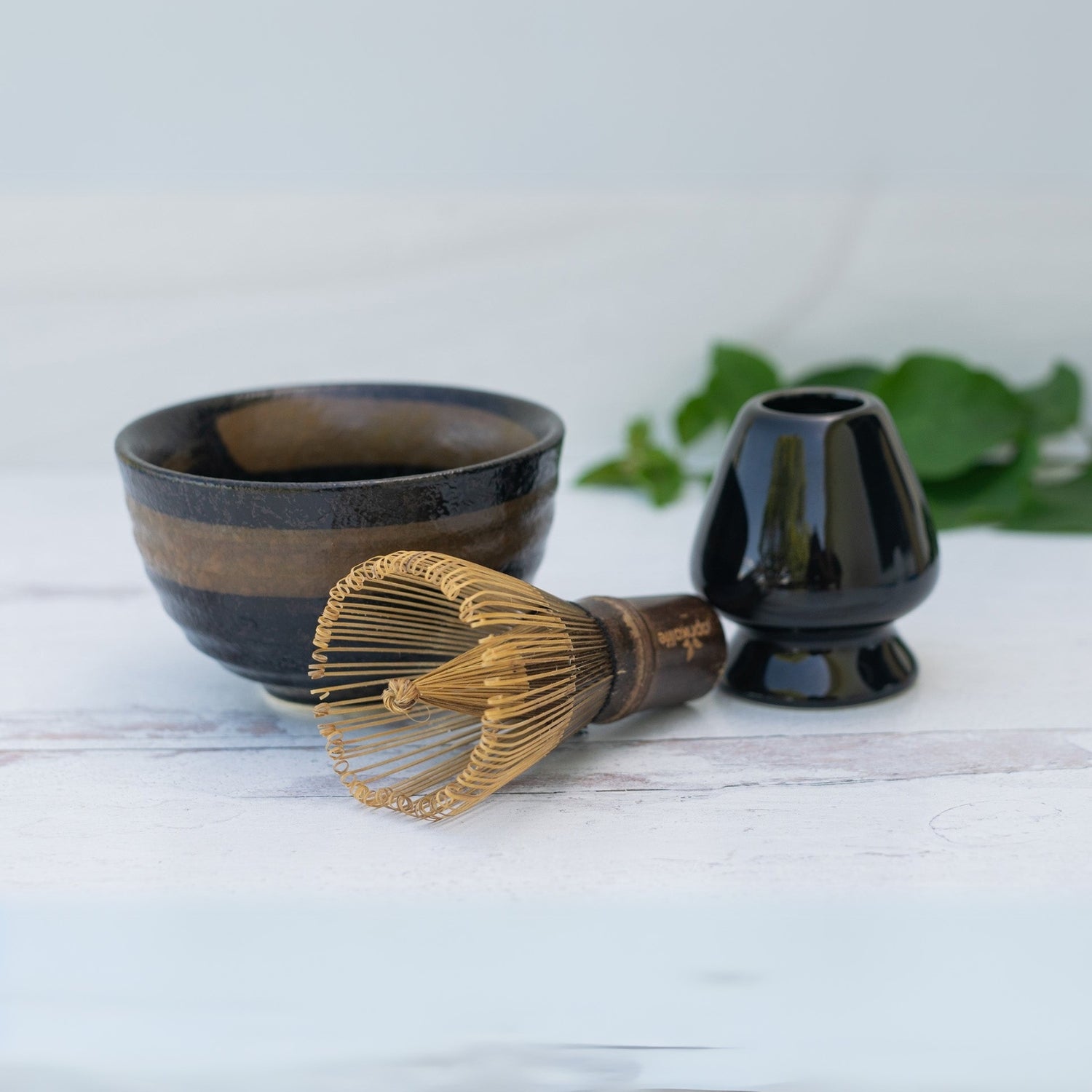 The Dark Brown Glaze Ceramic Matcha Bowl Set includes a bamboo whisk and holder, arranged elegantly on a white surface.
