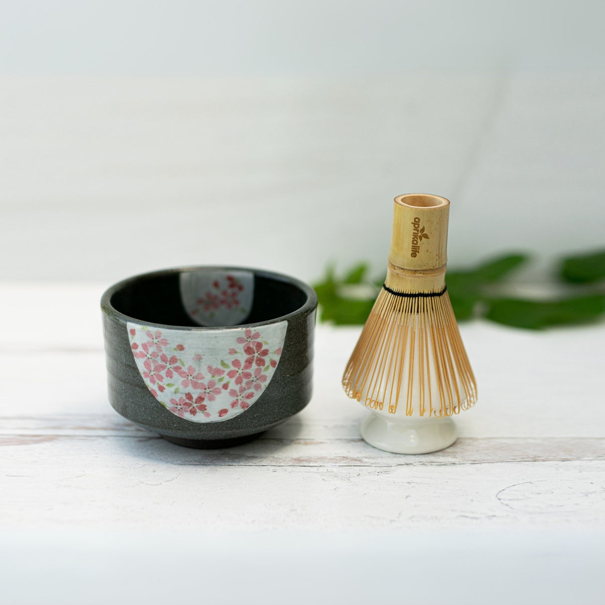 The Dark Grey Flowers Ceramic Matcha Bowl, showcasing Japanese pottery, beautifully complements an artisan bamboo tea whisk on a white surface.