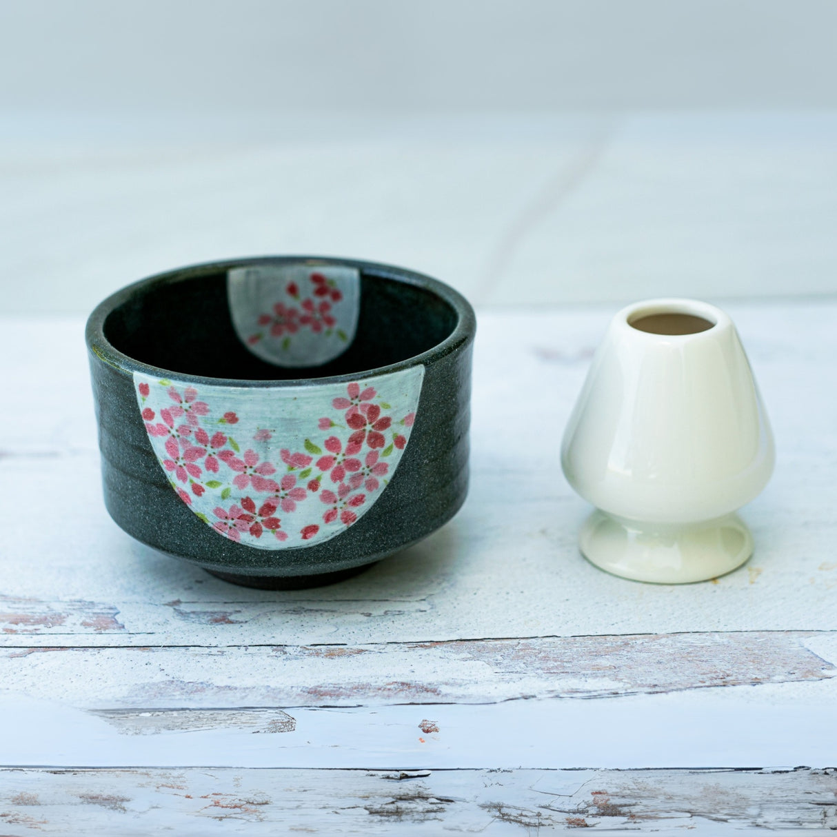A dark grey flowers ceramic matcha bowl, evoking traditional Japanese pottery, sits next to a small white vase on a light wooden surface.