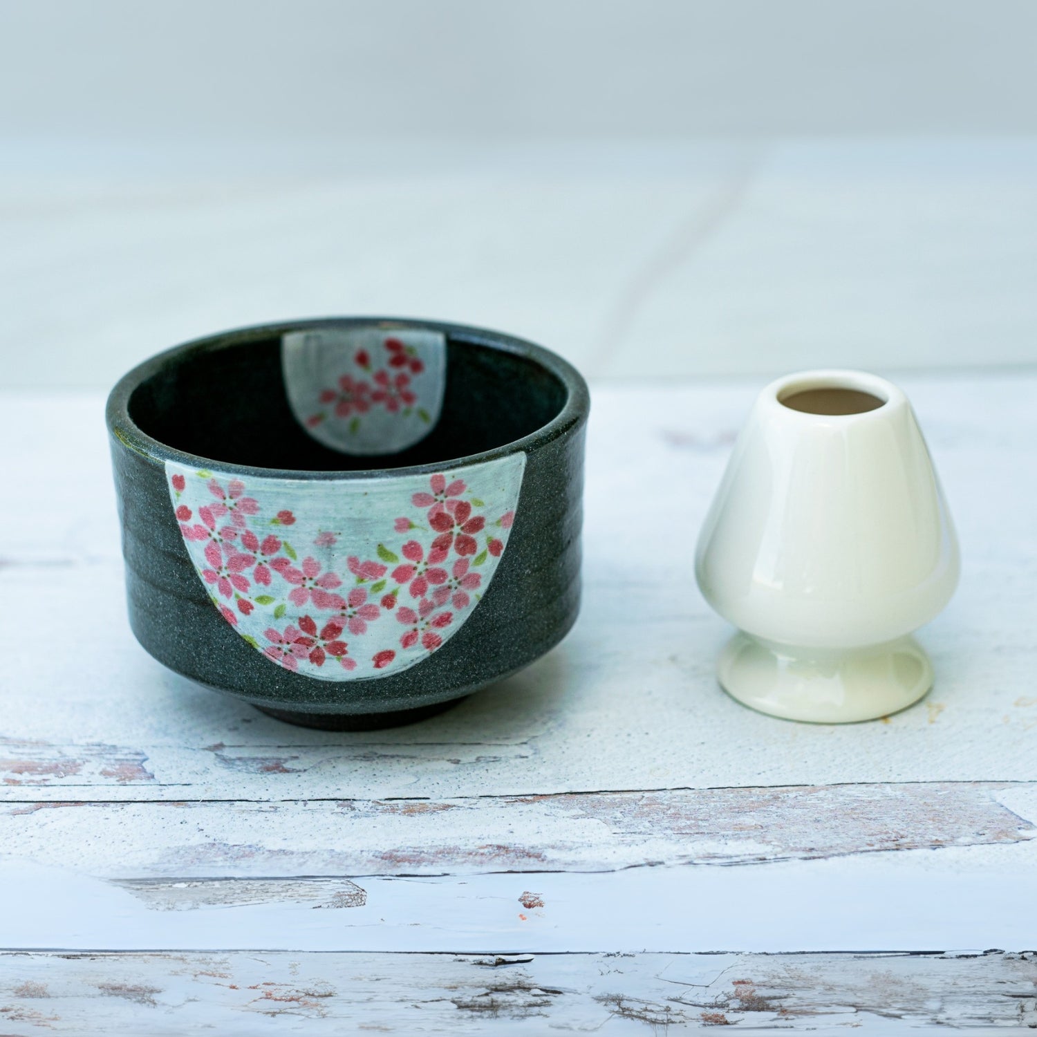 A dark grey flowers ceramic matcha bowl, evoking traditional Japanese pottery, sits next to a small white vase on a light wooden surface.