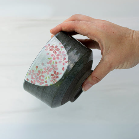 A hand cradles the Dark Grey Flowers Ceramic Matcha Bowl, showcasing Japanese elegance with its pink floral designs against a pristine white background.