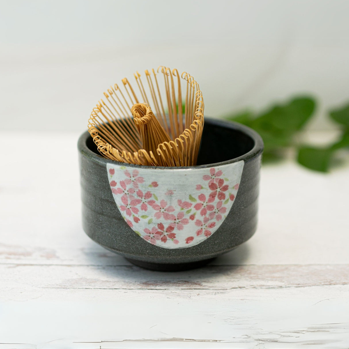 The Dark Grey Flowers Ceramic Matcha Bowl, with a bamboo matcha whisk and holder set, is shown on a white surface—a stylish pottery accessory for your matcha ritual.