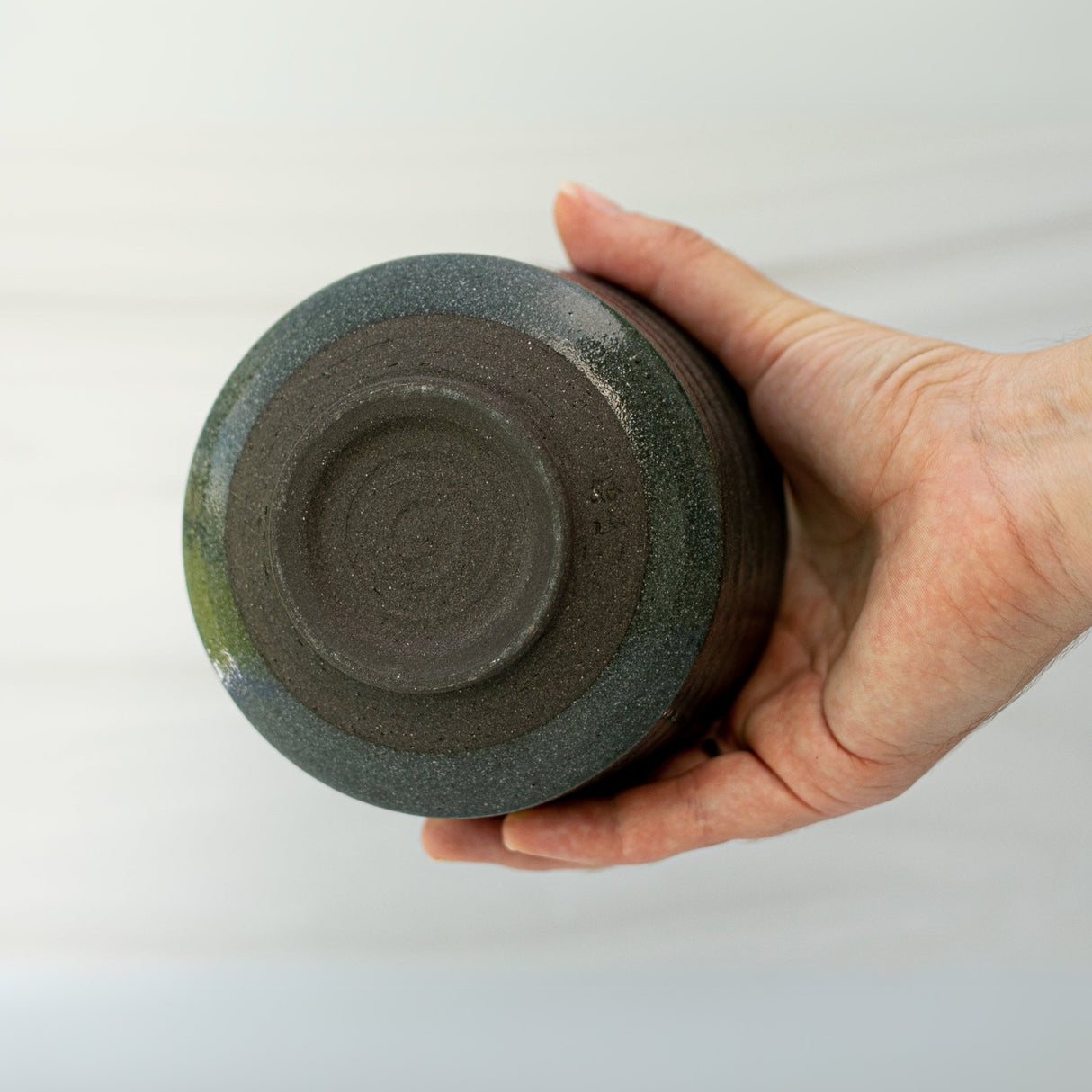 A hand holding the Dark Grey Flowers Ceramic Matcha Bowl, with its round base visible against a light background—an elegant showcase of Japanese design from our Matcha Bowl, Bamboo Whisk, and Whisk Holder Set.
