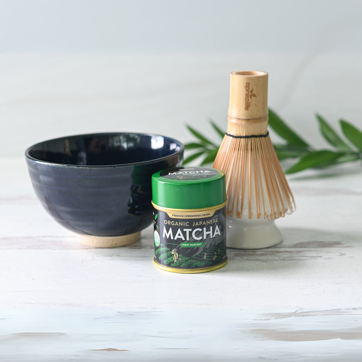 A Dark Navy Blue Ceramic Matcha Bowl with a bamboo matcha whisk and organic Japanese matcha rests elegantly on a white surface decorated with green leaves.