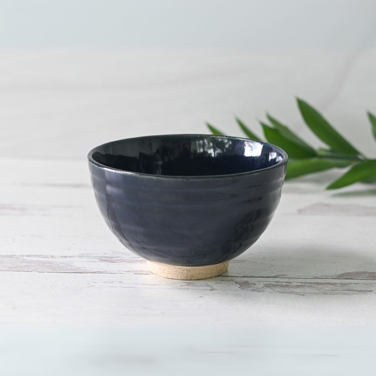 A Dark Navy Blue Ceramic Matcha Bowl from the Bamboo Matcha Whisk and Whisk Holder Set sits on a white surface, crafted with Minoyaki Pottery Techniques, with green leaves in the background.