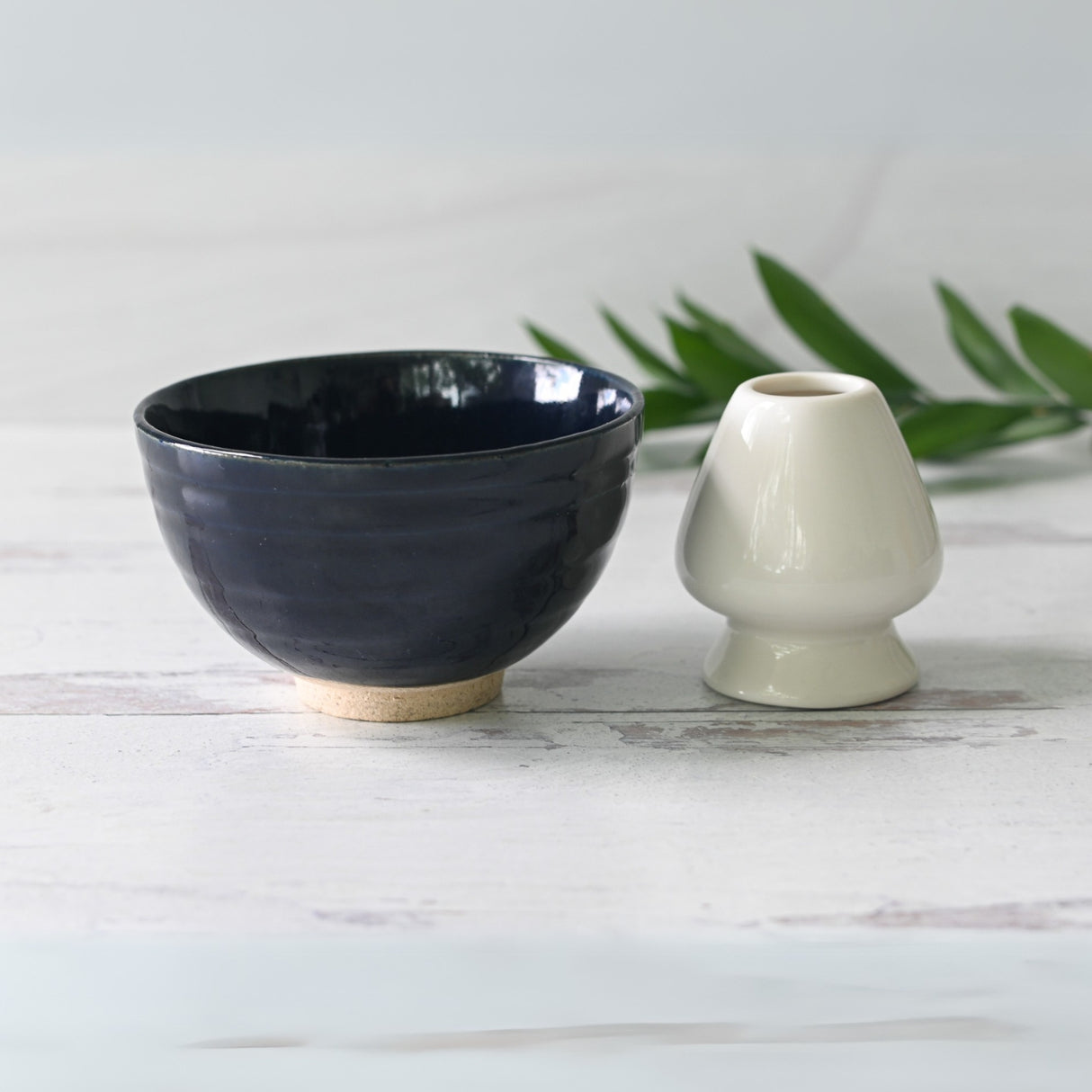 A Dark Navy Blue Ceramic Matcha Bowl elegantly sits alongside a white vase on a light wooden surface, with a lush green plant adding to the background's charm.