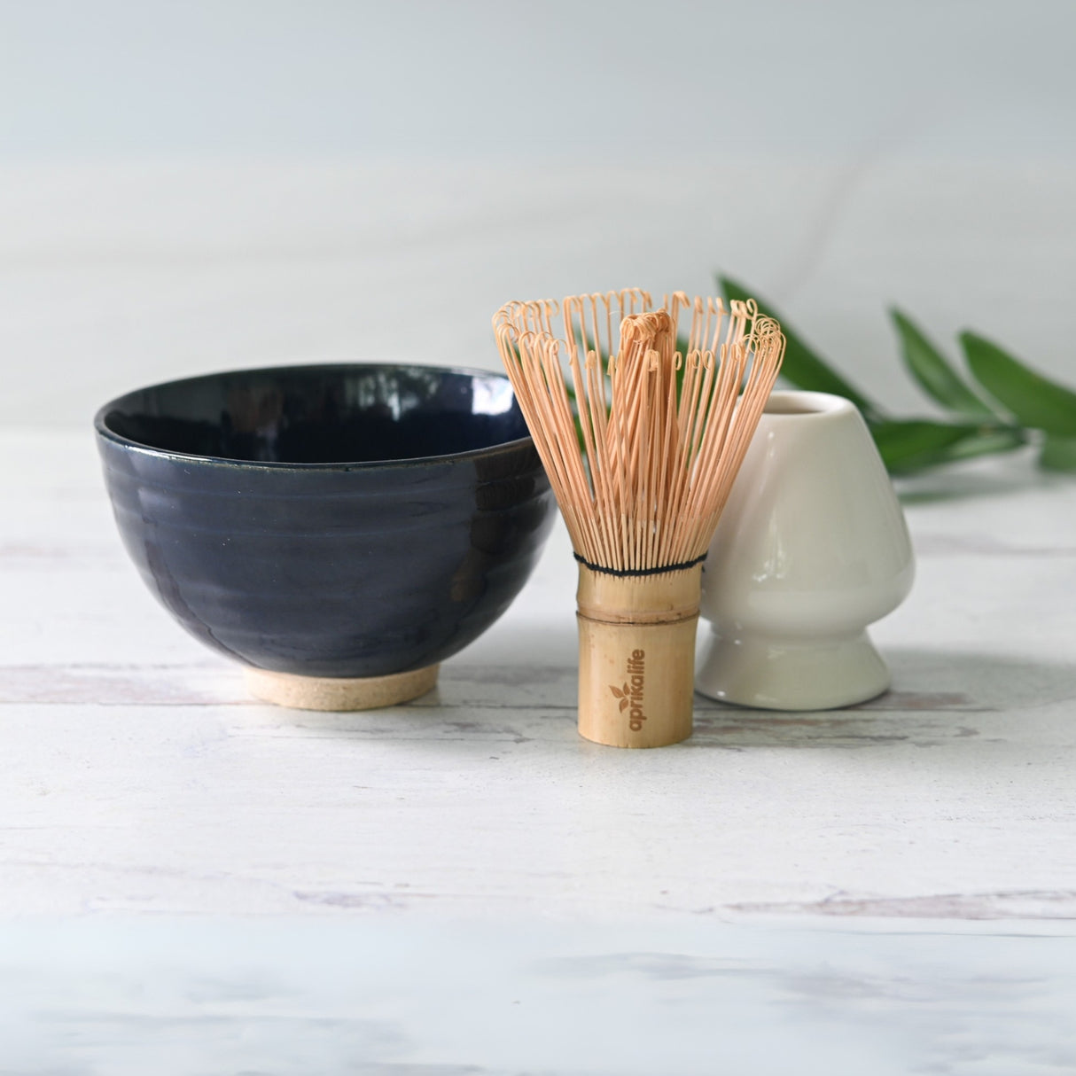 The Dark Navy Blue Ceramic Matcha Bowl, Bamboo Matcha Whisk, and Whisk Holder from the Organic Japanese Matcha Set rest elegantly on a light wooden surface with green leaves creating a serene backdrop.