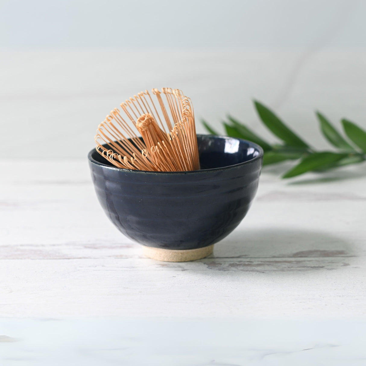 The Dark Navy Blue Ceramic Matcha Bowl holds a bamboo matcha whisk, elegantly placed on light wood with a green leaf in the background.