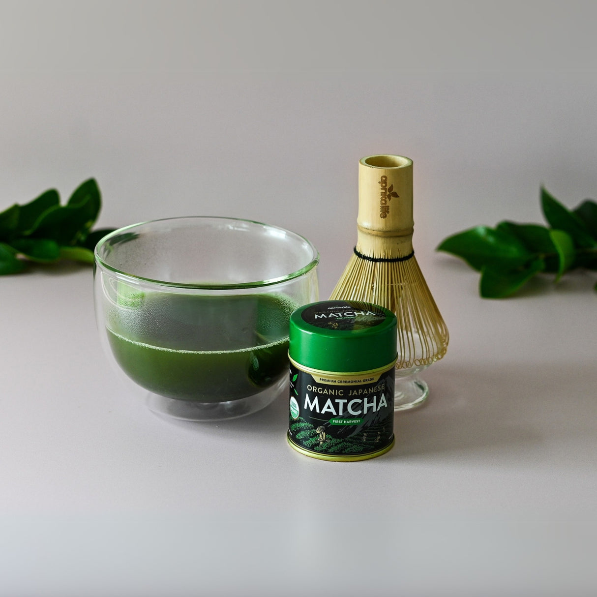 A Double Sided Glass Bowl holds a vibrant cup of matcha tea, paired with a bamboo whisk and a can of organic Japanese matcha, all set on a light surface adorned with green leaves.