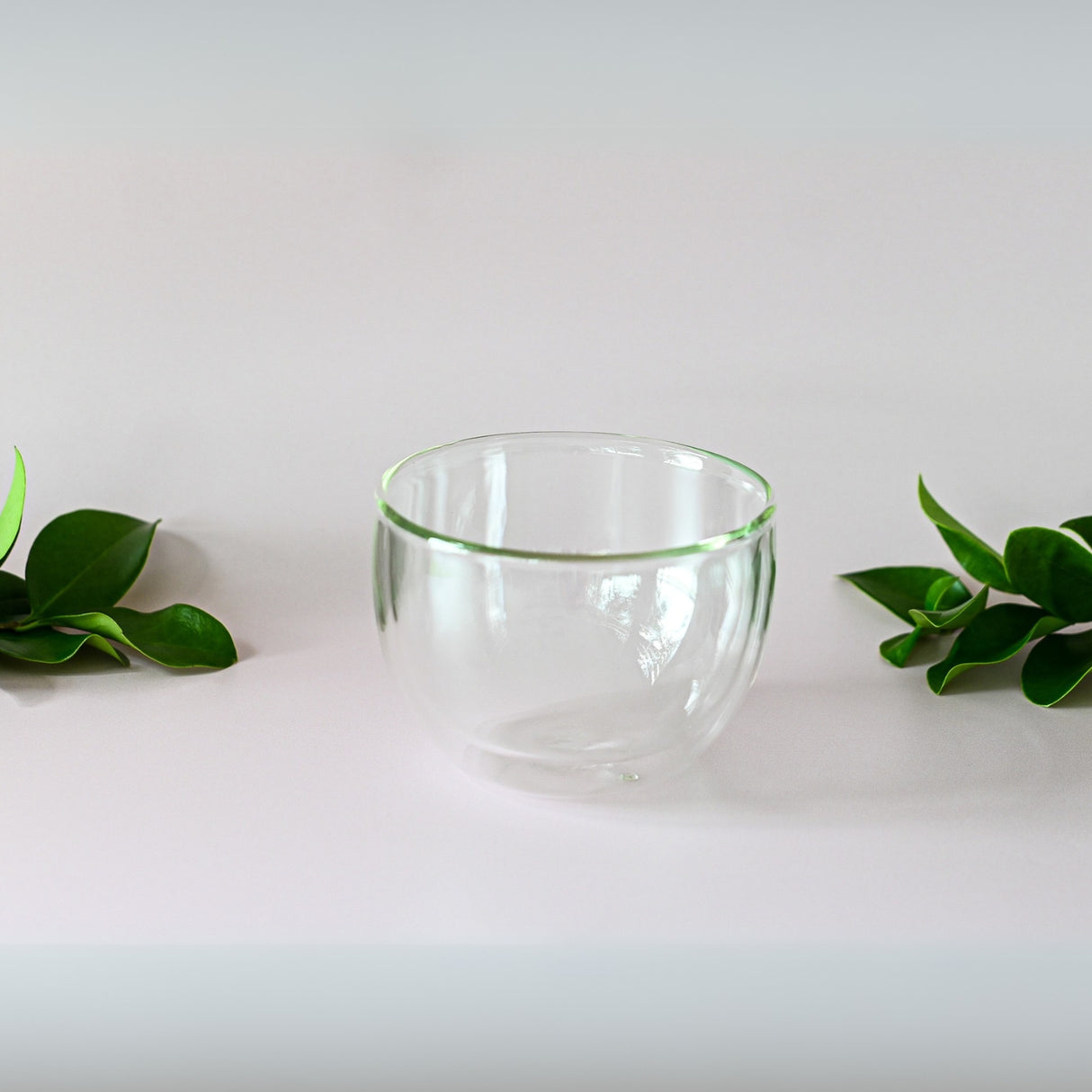 A Double Sided Glass Bowl sits on a white surface, surrounded by vibrant green leaves that capture the essence of freshly brewed teas.