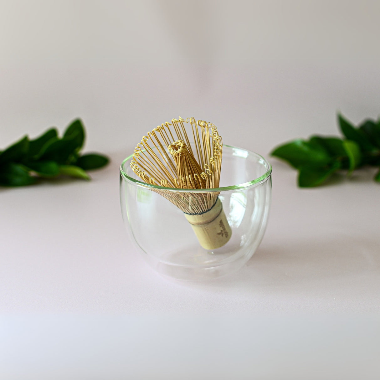 The Double Sided Glass Matcha Set features an elegant bamboo whisk nestled in a glass matcha bowl, framed by lush green leaves.