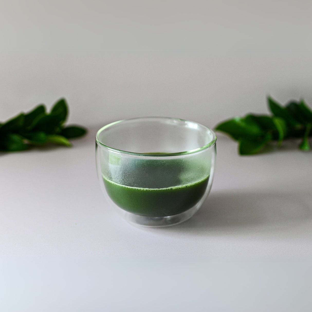 A Double Sided Glass Matcha Set, brimming with green juice and resembling a traditional matcha blend, rests on a light surface adorned with green leaves—the perfect setup for any Matcha Tea Starter Set fan.