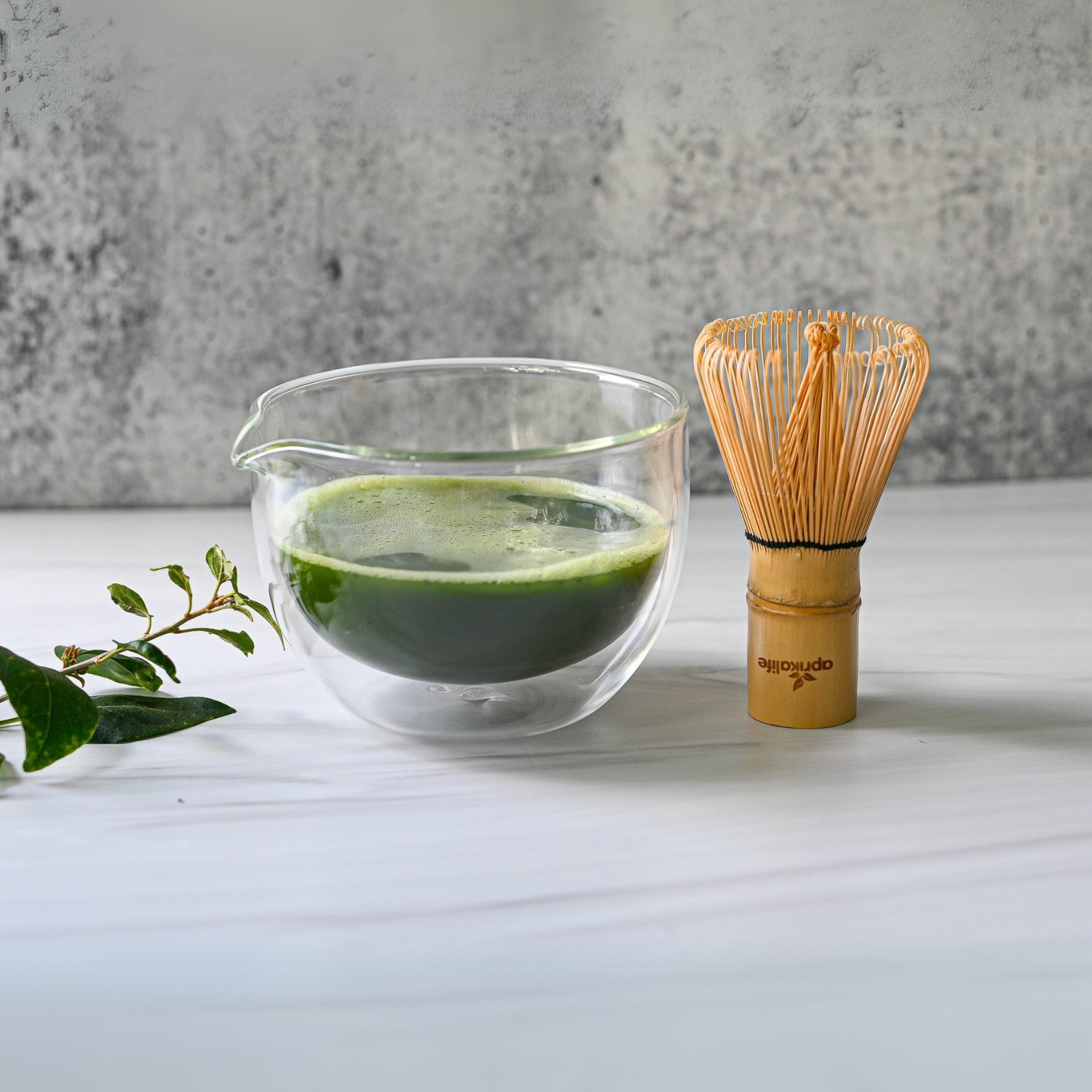 On a white surface sits a vibrant green matcha tea within a Double Sided (Double Wall) Glass Matcha Bowl with Spout, accompanied by a bamboo whisk and scattered leaves.