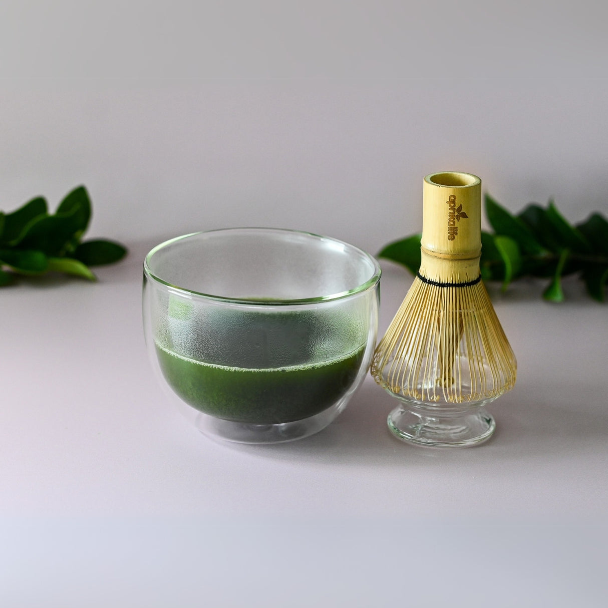 The vibrant green tea sits elegantly in the Double Sided Glass Bowl, with a bamboo whisk nearby, surrounded by lush green leaves.
