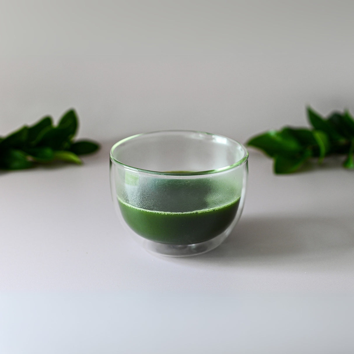 A Double Sided Glass Bowl filled with vibrant green liquid rests on a white surface, surrounded by lush green leaves in the background, capturing the tranquility of traditional tea ceremonies.