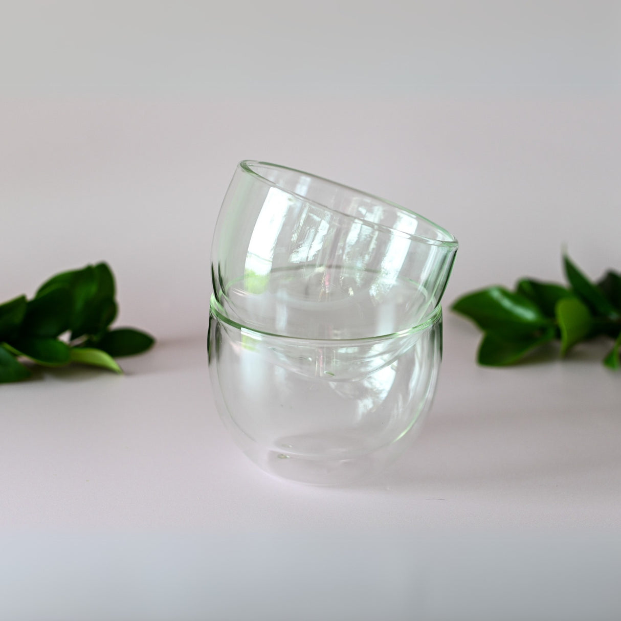 Two Double Sided Glass Bowls are stacked on a white surface, surrounded by green leaves, capturing the serene essence of matcha.