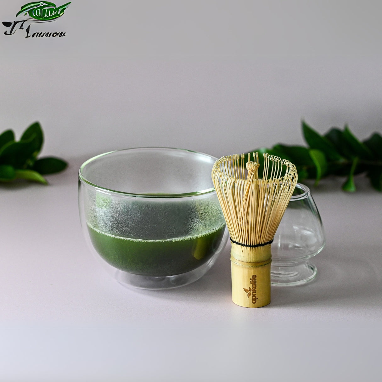 A Double Sided Glass Bowl elegantly sits on the table, filled with vibrant matcha tea. A bamboo whisk lies nearby, surrounded by lush green leaves, capturing the essence of quality green teas in a serene setting.