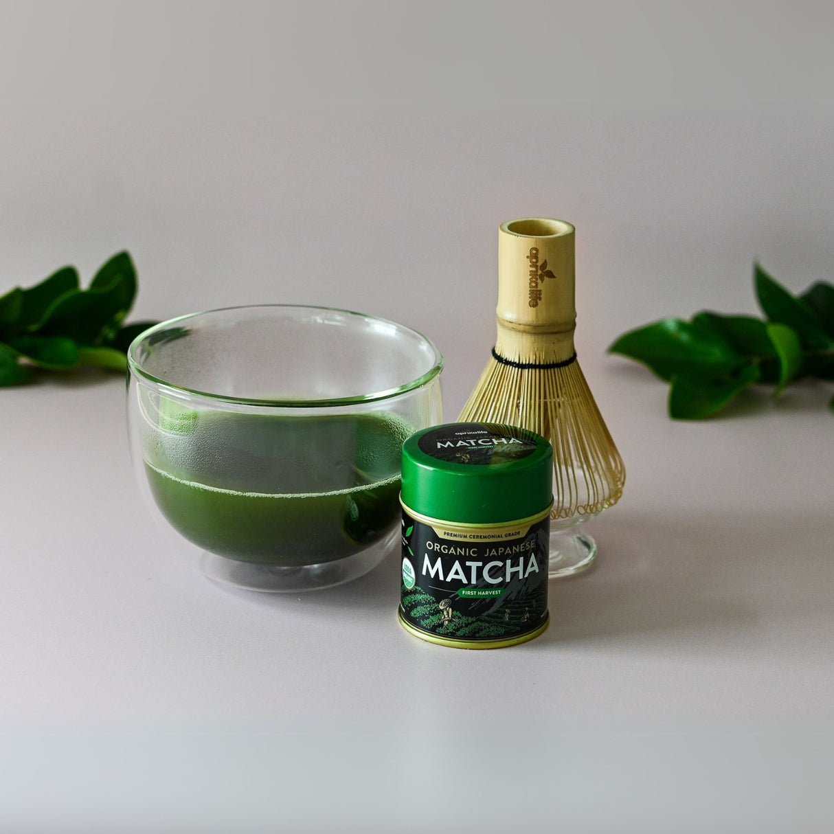 The Double Sided Glass Matcha Set, featuring a vibrant matcha bowl, tin, and bamboo whisk, is elegantly arranged on a white surface with lush green leaves.