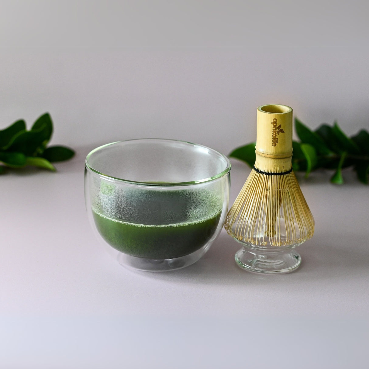 The Double Sided Glass Matcha Set includes an elegant clear glass bowl of matcha tea beside a bamboo whisk, complemented by lush green leaves for a serene backdrop.