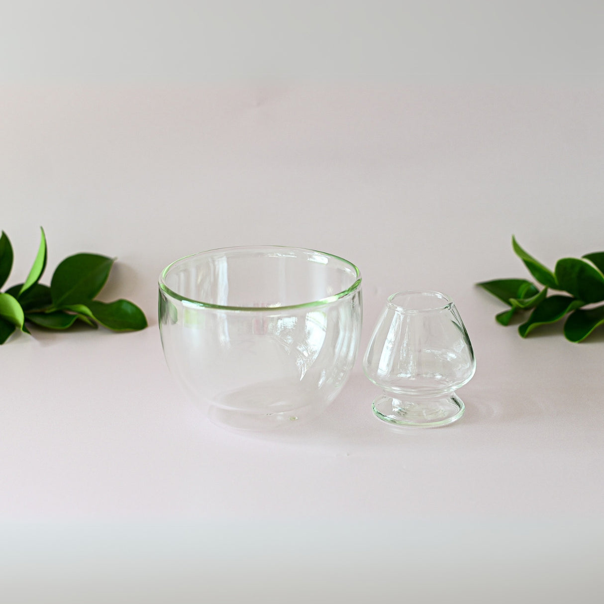 Two clear bowls from the Double Sided Glass Matcha Set sit on a white surface with green leaves, paired with a bamboo whisk—ideal for any matcha starter collection.