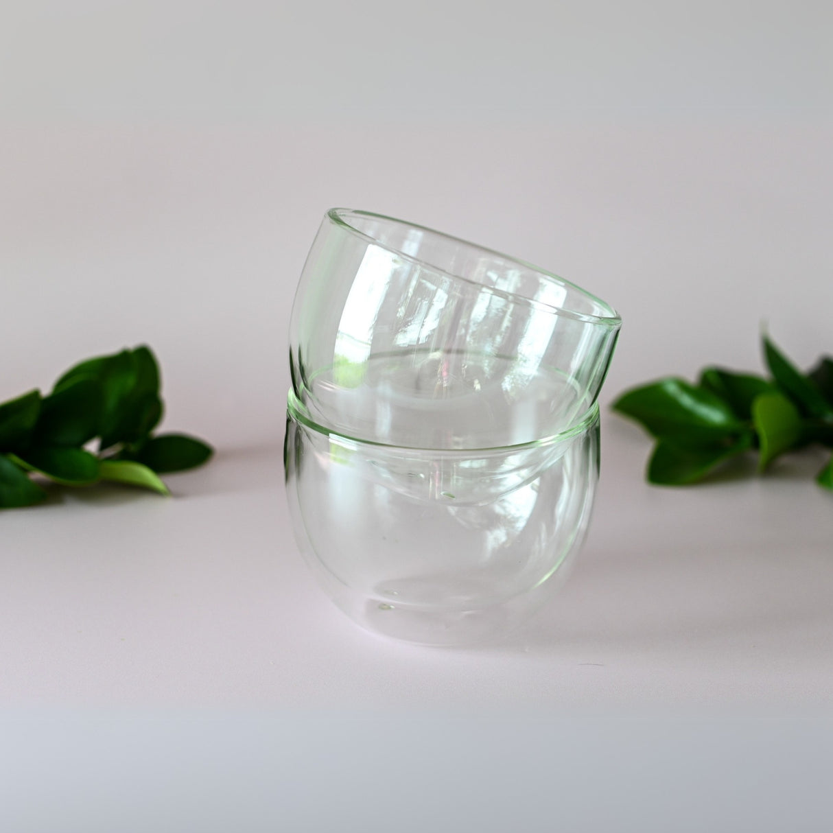 Two clear cups from the Double Sided Glass Matcha Set are stacked amid lush green leaves on a pristine white surface, creating an ideal setting.