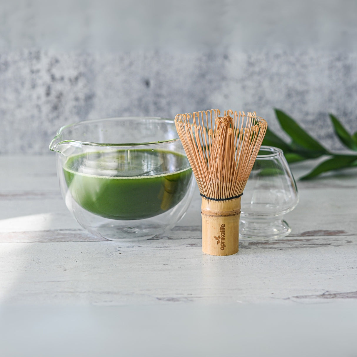 Matcha tea in a Double Sided Glass Matcha Bowl with Spout, accompanied by a bamboo whisk on a white wooden surface, surrounded by greenery. This setup showcases the elegant simplicity and cultural richness of green teas.