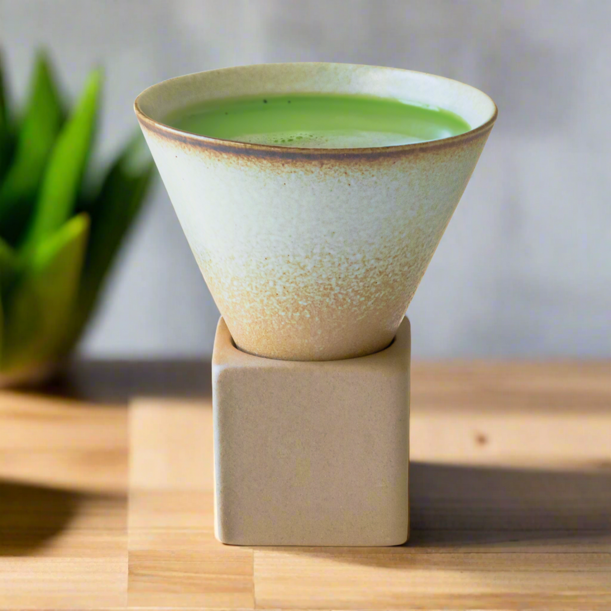 A Brown Coffee Mug with Base - 6.8 oz rests elegantly on a wooden surface, filled with green tea, and a plant's blurred silhouette graces the background.