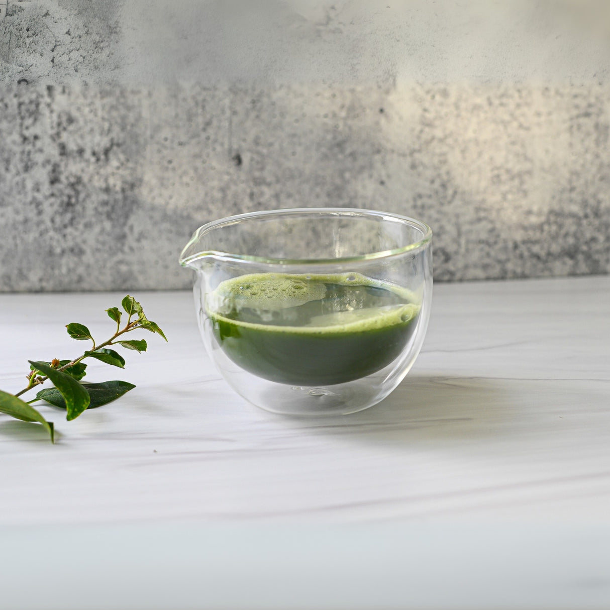 A Double Sided (Double Wall) Glass Matcha Bowl with Spout, surrounded by a leafy branch on a white surface, embodies the tranquility of traditional tea preparation.