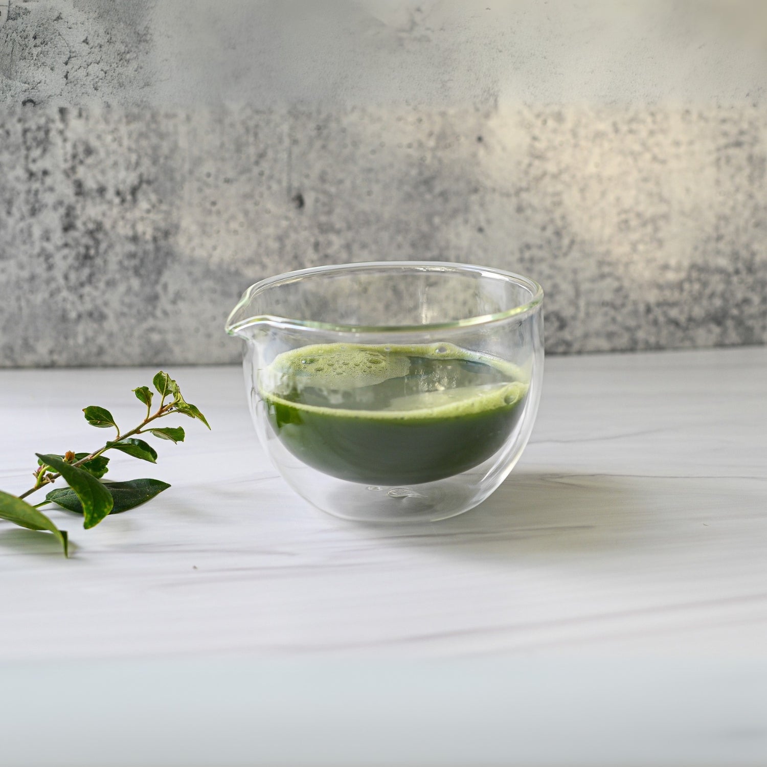 A Double Sided (Double Wall) Glass Matcha Bowl with Spout, surrounded by a leafy branch on a white surface, embodies the tranquility of traditional tea preparation.