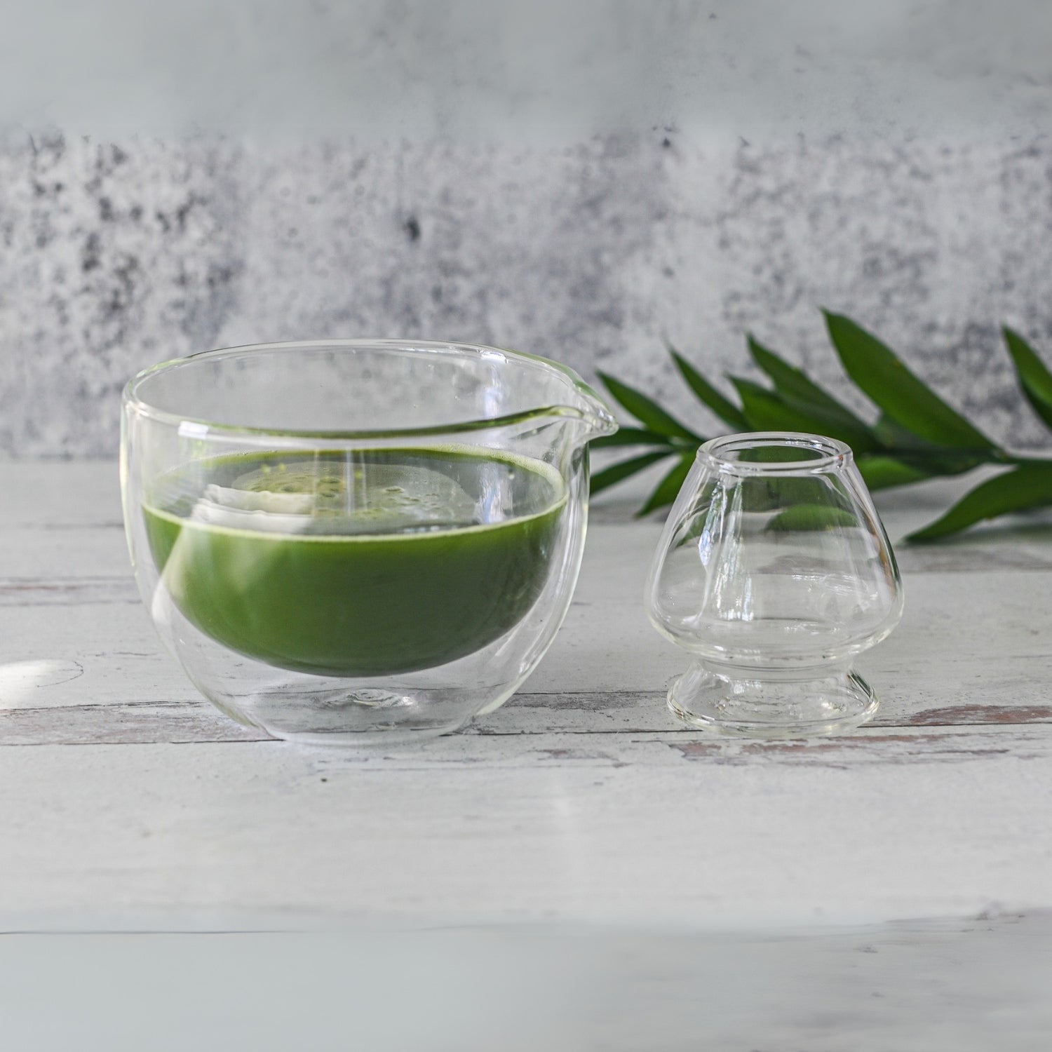 A Double Sided Glass Matcha Bowl with vibrant green liquid sits beside an empty glass on a white wooden surface, accented by a green leaf. This serene scene is perfect for using the Bamboo Matcha Whisk and Holder from the Double Wall Matcha Tea Set.