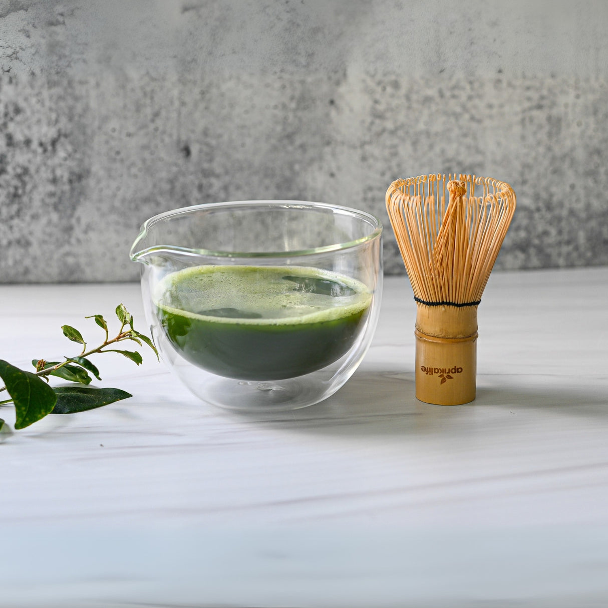 A Double Sided Glass Matcha Bowl with Spout, filled with vibrant tea, sits elegantly beside a Bamboo Matcha Whisk and a small green branch on a pristine white surface.