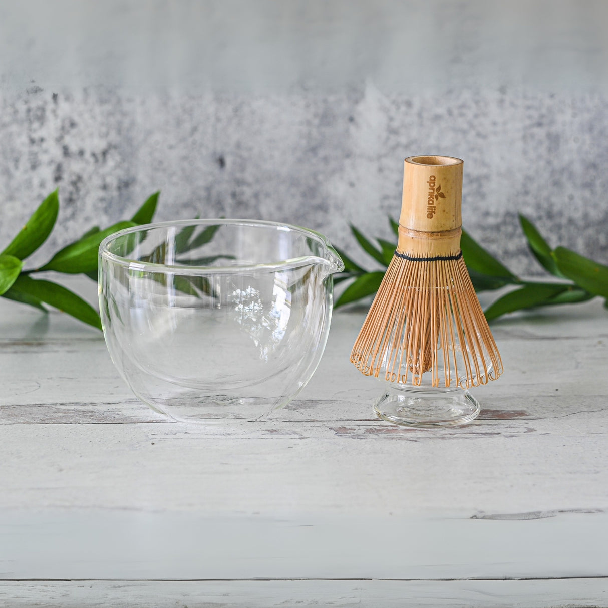 A Double Sided Glass Matcha Bowl with Spout and Bamboo Matcha Whisk rest elegantly on a light wooden surface, framed by lush green leaves.