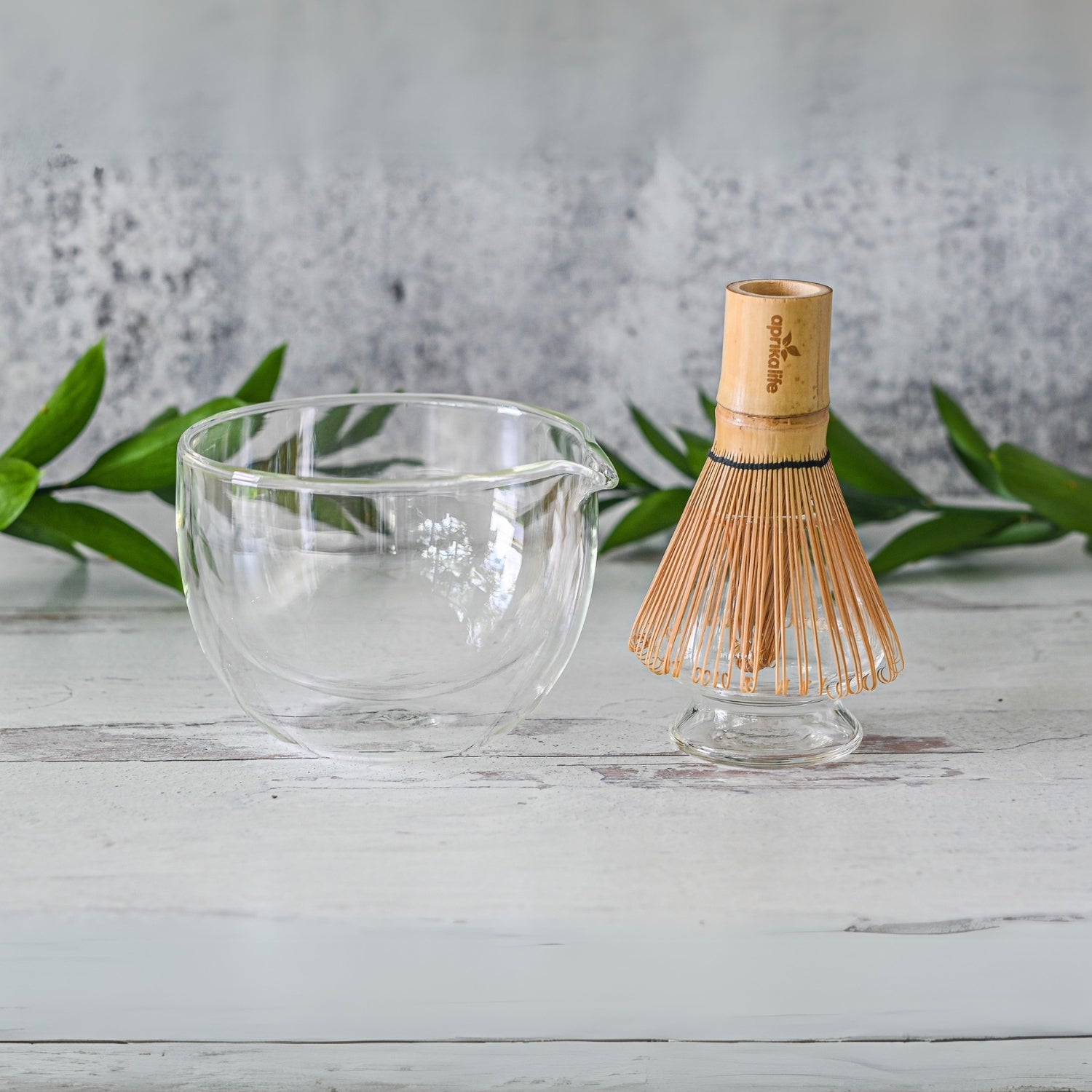 A Double Sided Glass Matcha Bowl with Spout and Bamboo Matcha Whisk rest elegantly on a light wooden surface, framed by lush green leaves.