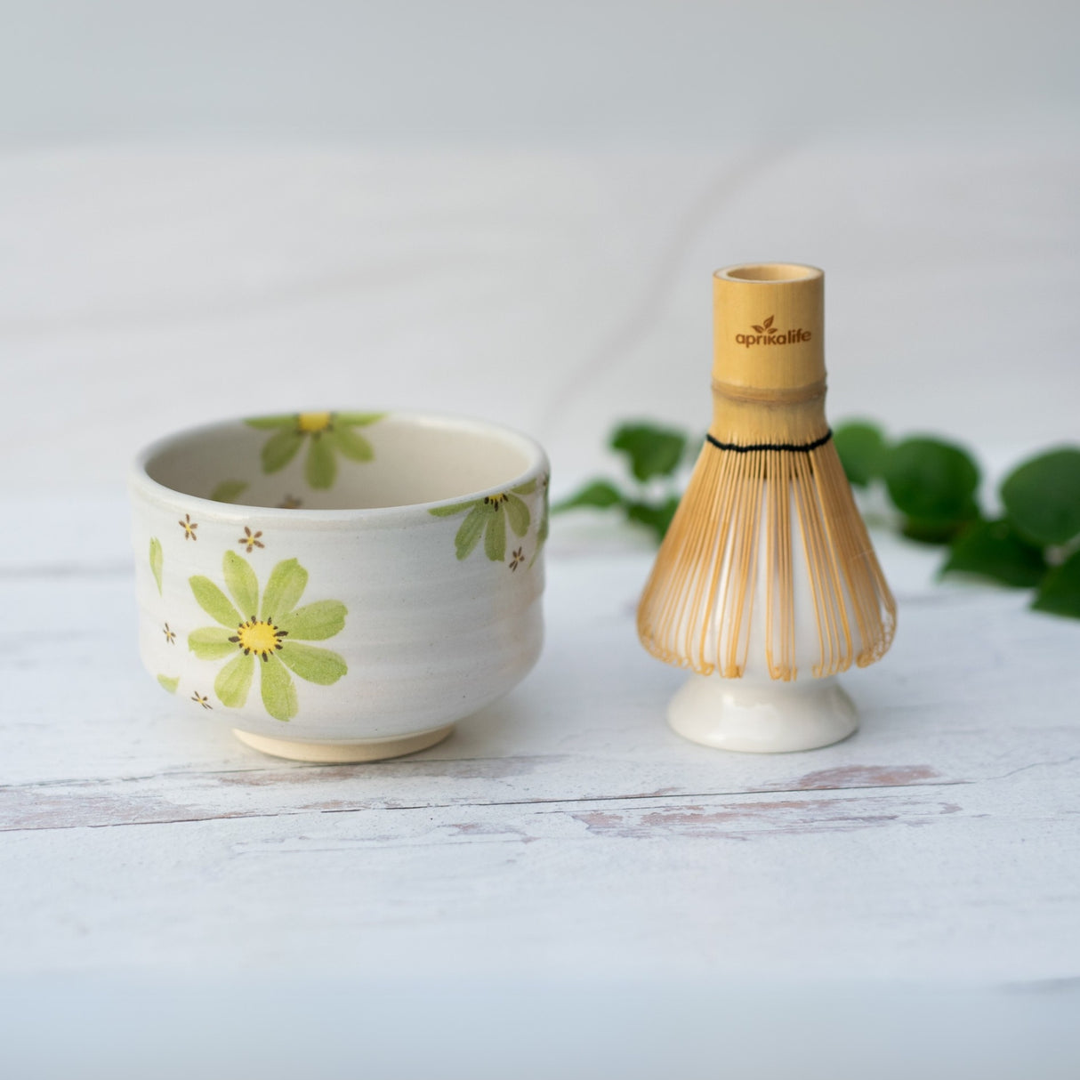 The Green Flowers Ceramic Matcha Bowl, Bamboo Matcha Whisk, and Whisk Holder Set features a floral Minoyaki bowl and bamboo whisk—arranged on a white wooden surface with green leaves—perfect as your matcha starter set.