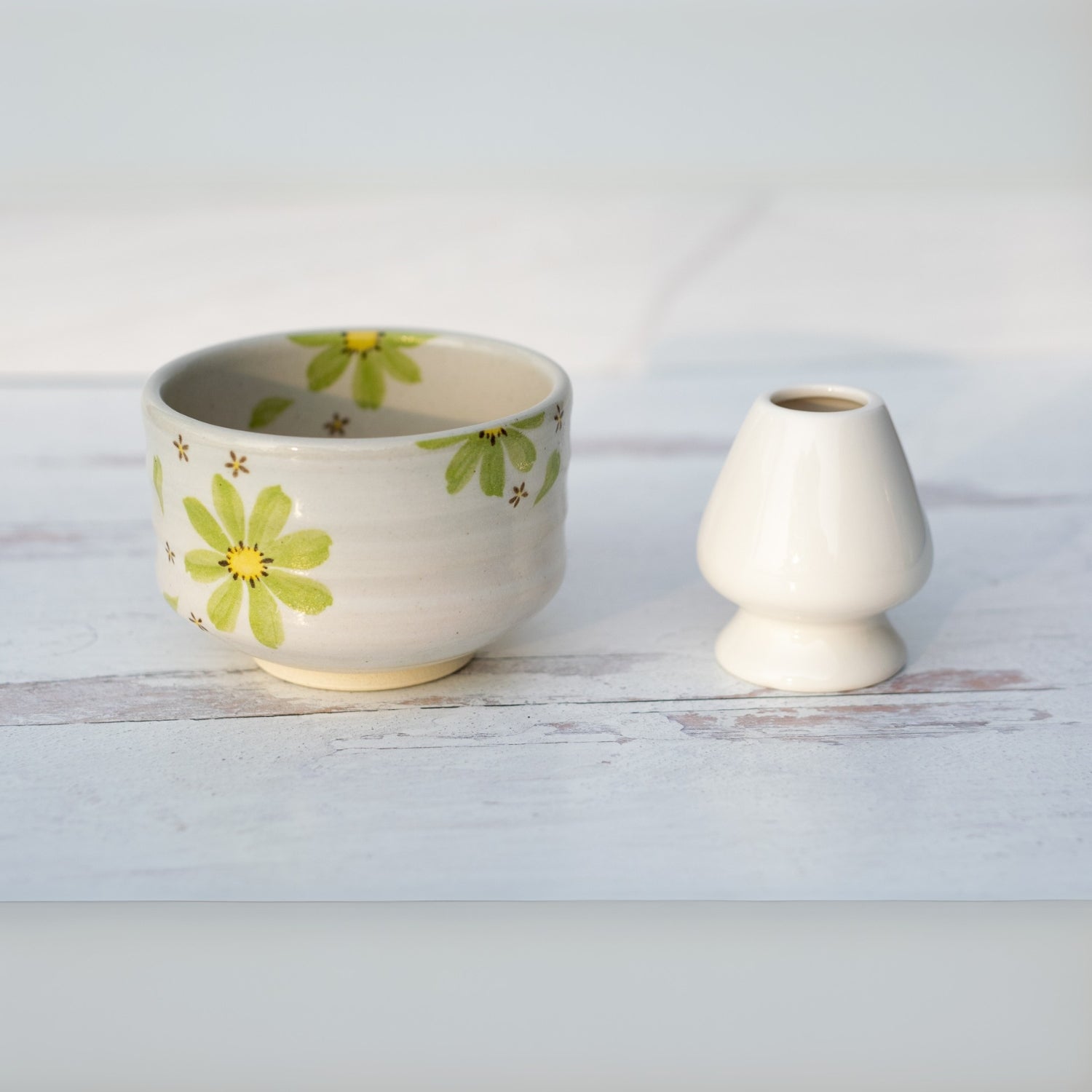 A Green Flowers Ceramic Matcha Bowl from the Matcha Set, featuring a floral design, sits beside a plain white vase on a light surface, inspired by traditional Japanese pottery.