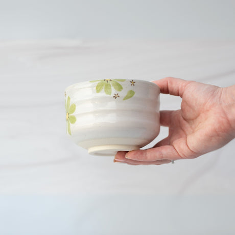 A hand holds a white ceramic bowl with green floral patterns from the Green Flowers Ceramic Matcha Bowl, Bamboo Matcha Whisk, Whisk Holder and Organic Japanese Matcha Set against a light background.